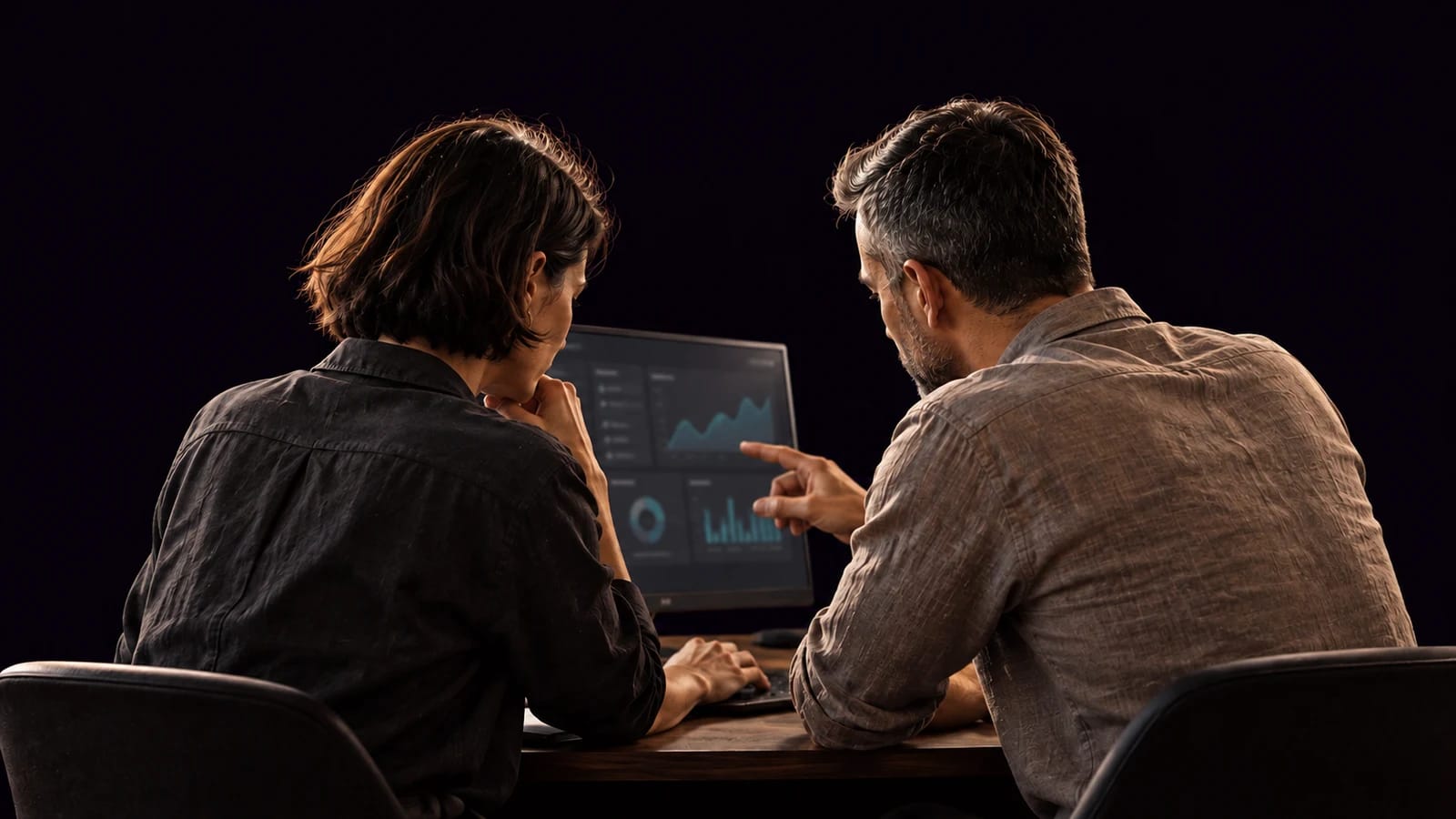 Two professionals seen from behind reviewing a screen, cut out against a dark background