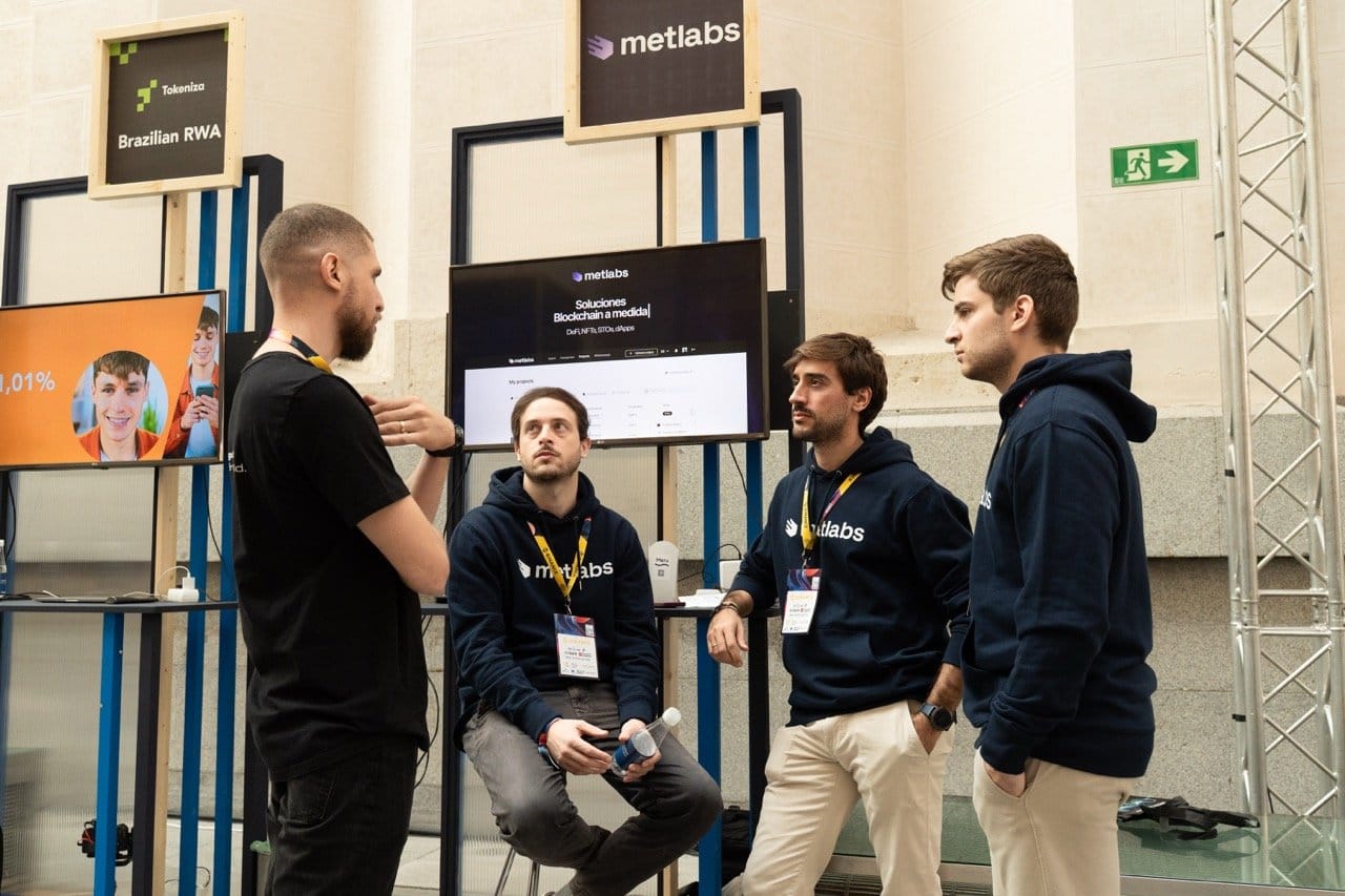 Metlabs team talking at their Merge Madrid stand next to the product screen