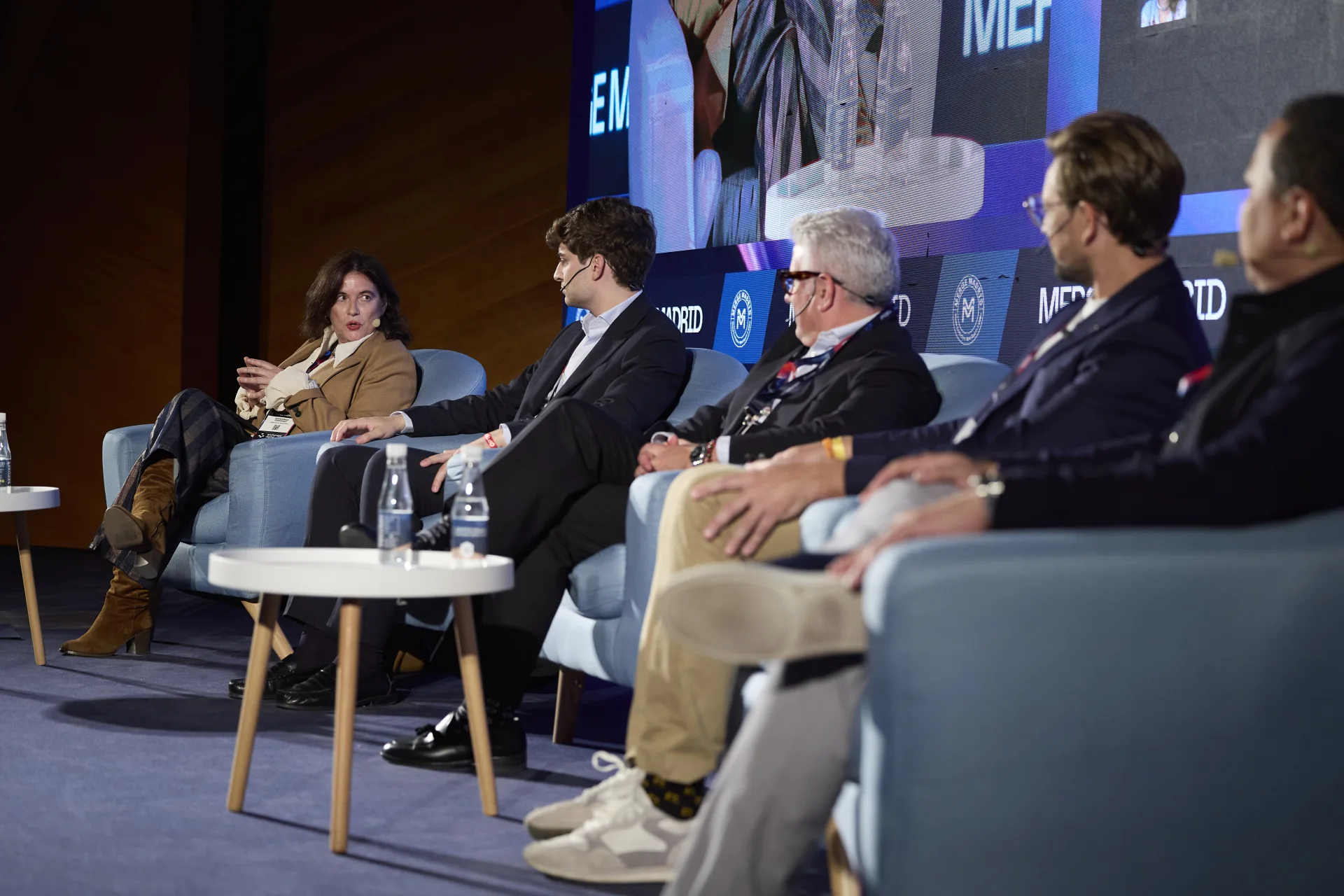 Covadonga Fernández moderating the MERGE Stage panel, seen from the side with the five participants in their armchairs
